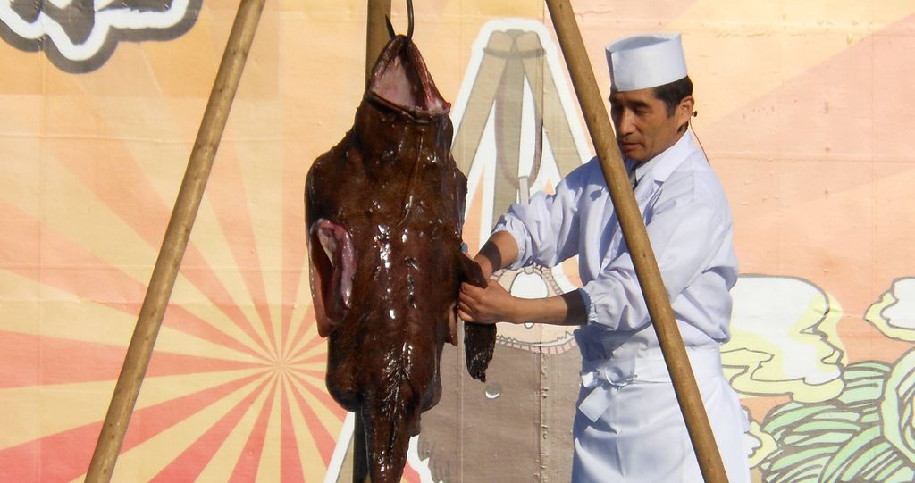 Cook in classical attire hanging and cutting an anglerfish during a live demonstration at the Oarai Anko Festival, with a colorful mural in the background.
