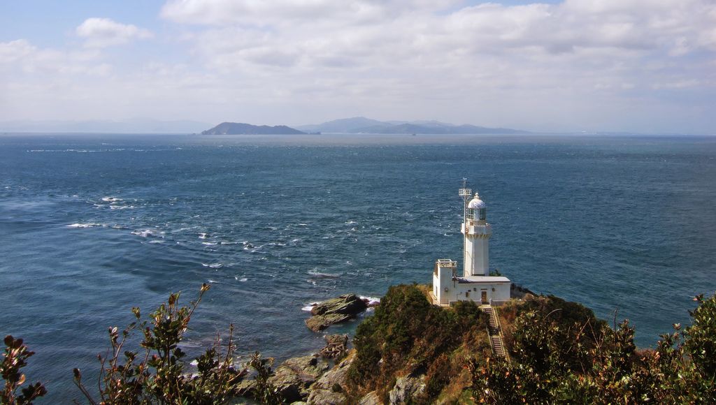 Ein weißer Leuchtturm steht auf einer felsigen Klippe mit Blick auf die Bungo-Straße in Japan, mit dem weiten blauen Ozean, der sich unter einem teilweise bewölkten Himmel bis zum Horizont erstreckt. In der Ferne sind schwach Inseln zu erkennen.