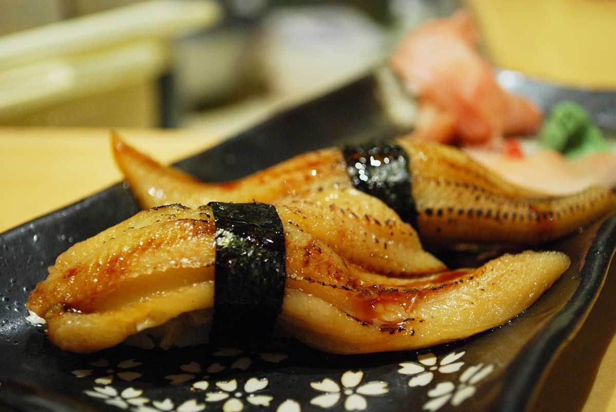 Two pieces of nigiri sushi with grilled eel, wrapped in nori, on a decorative black plate, accompanied by pickled ginger and wasabi