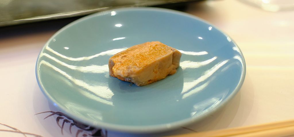 A piece of Ankimo, monkfish liver, delicately placed on a light blue sushi plate, highlighted by the simple elegance of its presentation.