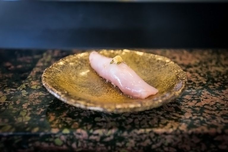 Einzelnes Stück Albacore-Sushi Nigiri, kunstvoll platziert auf einer goldfarbenen Keramikplatte über einer Marmoroberfläche.