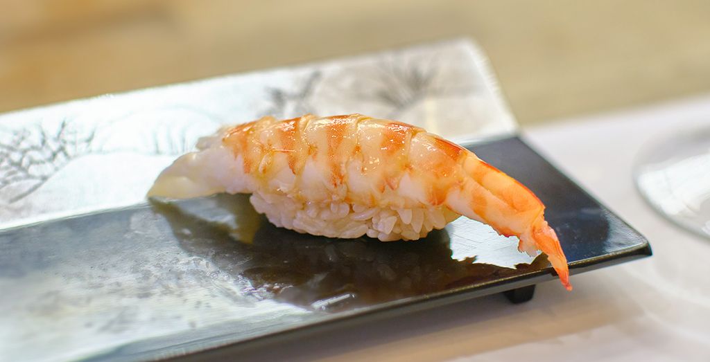 Ein Stück Ebi Nigiri Sushi, bestehend aus einer geschälten Garnele auf einer kleinen Portion Reis, serviert auf einem rechteckigen Keramikteller mit einer dezenten Bemalung im Hintergrund.