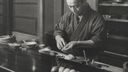 Schwarzweißfotografie. Ein Mann in dunkler Baumwoll-Arbeitskleidung an einer polierten Holztheke, mit gewölbten Händen, die gerade ein Nigiri formen. Kleine Schalen, ein Pinsel und ein Glas stehen auf der Theke; einige fertige Nigiri-Stücke sind vorn am Rand zu sehen. Traditionell japanisches Interieur im Hintergrund.
