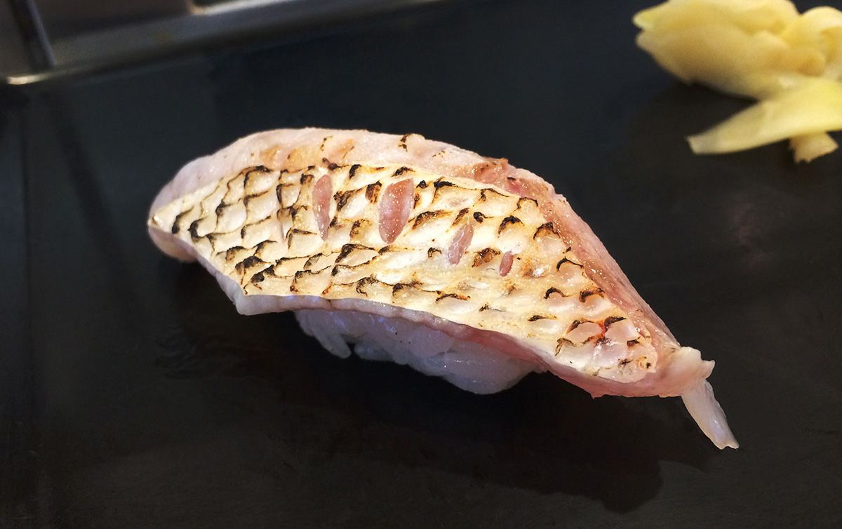 Aburi-Stil Nigiri-Sushi mit gegrillter Fischhaut auf einem schwarzen Keramikteller, begleitet von eingelegtem Ingwer am Rand