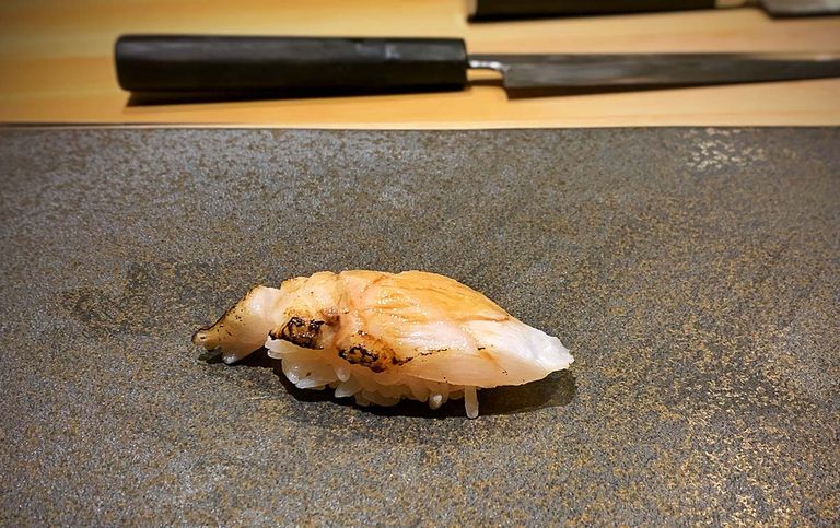 Einzelnes Nigiri-Sushi mit leicht angebrannter Nodoguro-Fischauflage auf einem dunklen Steinplatten-Tisch, mit unscharfem traditionellem japanischem Messer im Hintergrund