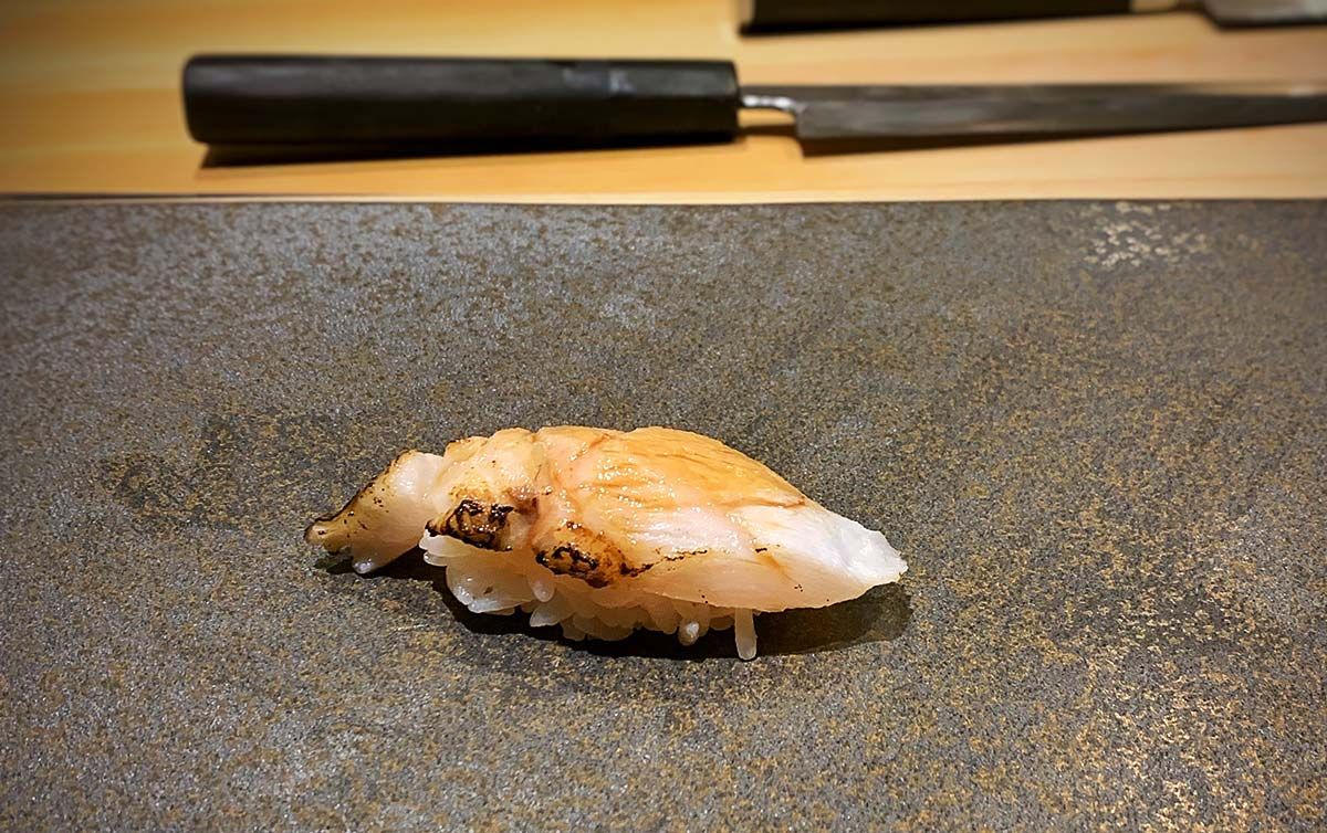 Einzelnes Nigiri-Sushi mit leicht angebrannter Nodoguro-Fischauflage auf einem dunklen Steinplatten-Tisch, mit unscharfem traditionellem japanischem Messer im Hintergrund