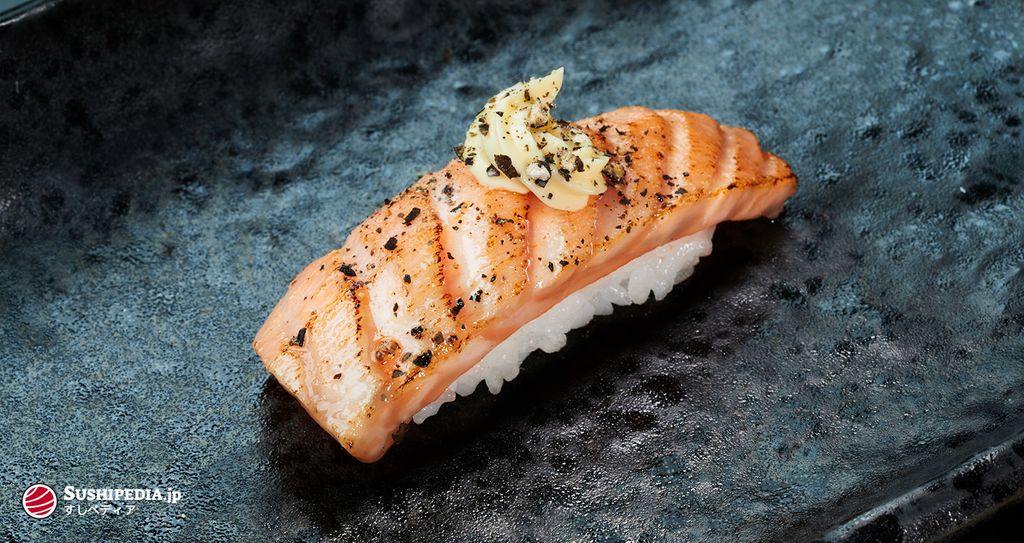 Seared Sake Lachs Nigiri, kunstvoll angerichtet mit einer Scheibe Butter und schwarzen Sesamsamen auf dunklem Untergrund.
