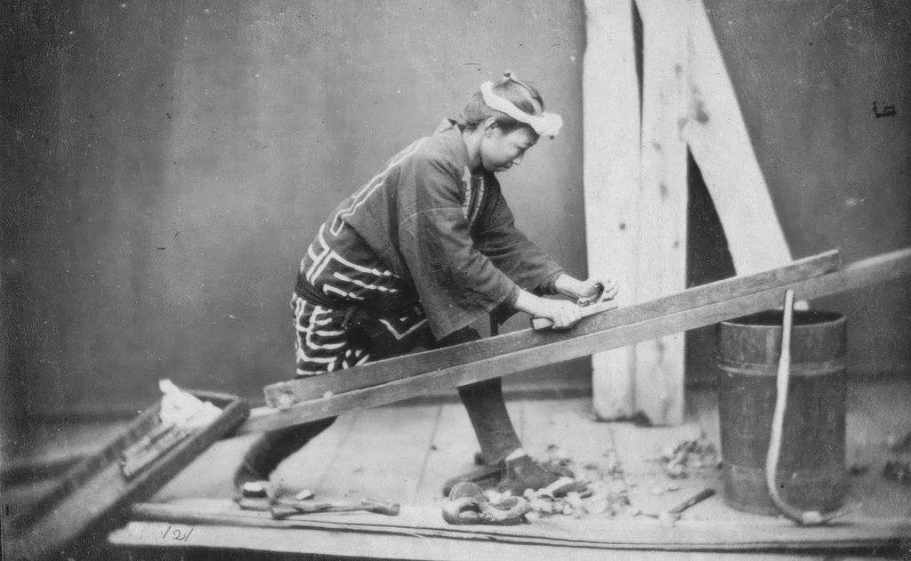 Historical black-and-white photograph of a Japanese carpenter planing a wooden beam. He wears a headband and traditional work clothing; wood shavings and hand tools are scattered on the floor.