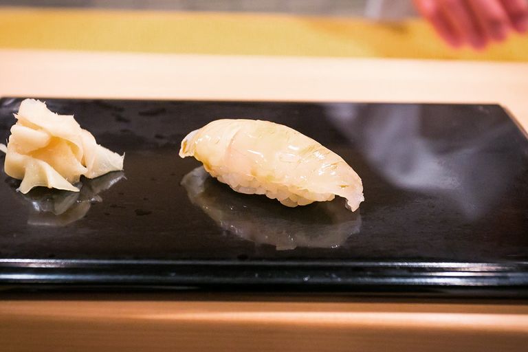 Das Bild zeigt ein einzelnes Stück Heilbutt Sushi Nigiri auf einem glänzenden schwarzen Teller mit eingelegtem Ingwer an der Seite. Der Sushi Reis ist gerade unter der durchscheinenden Fischscheibe sichtbar, was die kunstvolle Balance der Handwerkskunst des Sushi-Kochs hervorhebt.