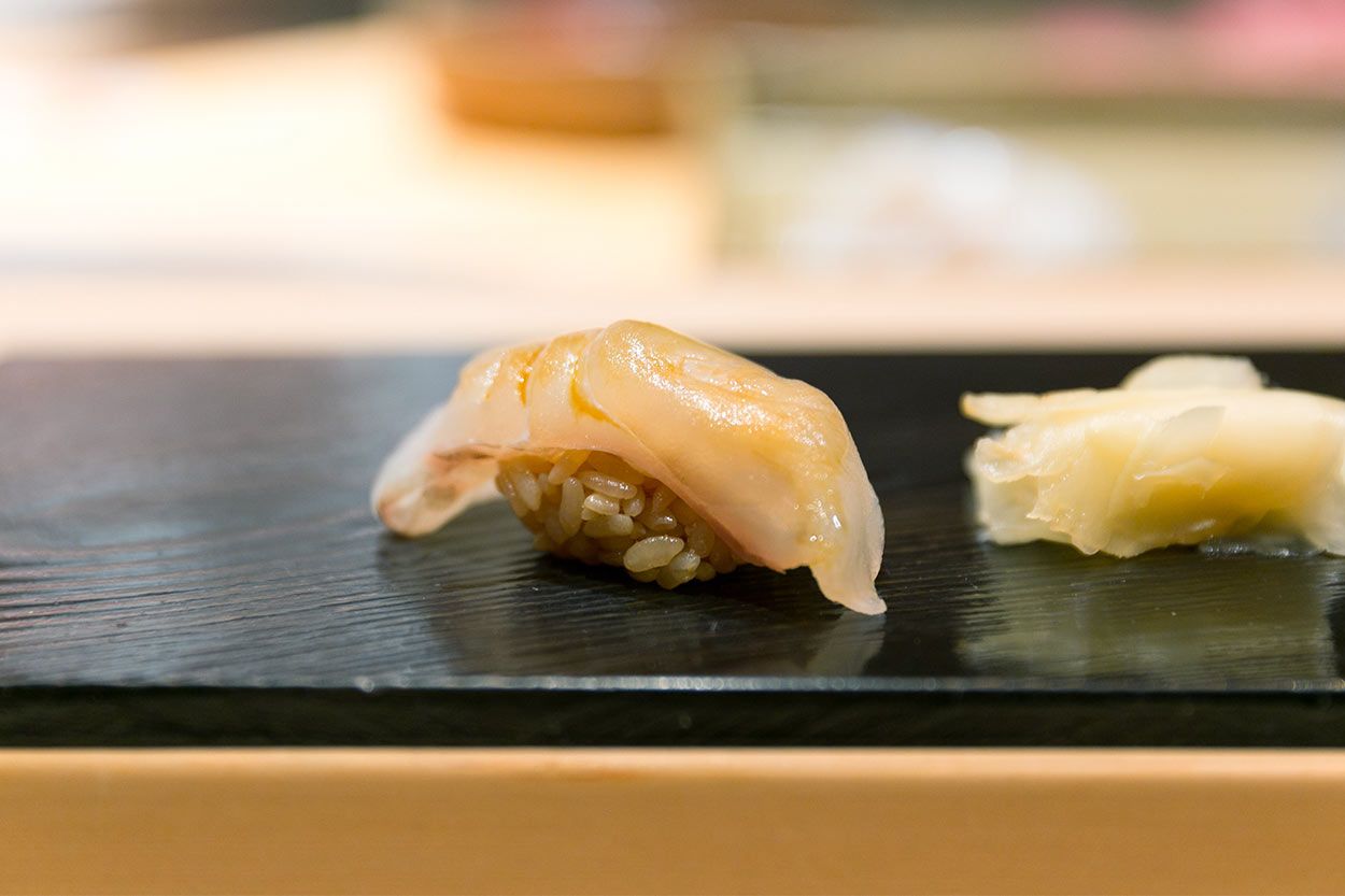 Ein Bild, das ein einzelnes Stück Heilbutt Nigiri Sushi präsentiert, mit einer dünnen, durchscheinenden Scheibe Heilbutt, die elegant über einem Haufen gewürzten Reises drapiert ist, serviert auf einem dunklen, minimalistischen Sushi-Teller mit einem unscharfen Hintergrund, der die Einfachheit und Konzentration auf das Sushi selbst hervorhebt.