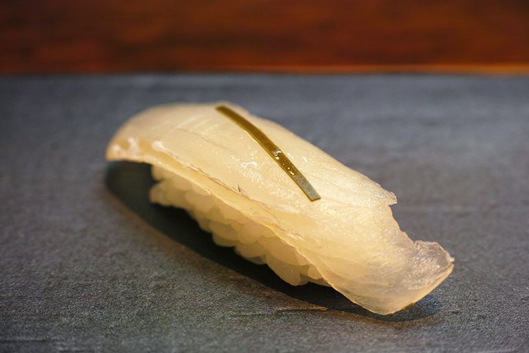 Eine Nahaufnahme von einem zubereiteten Hamachi Nigiri Sushi. Das Sushi zeigt eine Scheibe glänzenden, blassgelben Hamachi-Fisch, sanft auf einem Bett aus Sushi-Reis platziert, akzentuiert mit einem dünnen Streifen Kombu-Alge oben drauf, um den Umami-Geschmack zu verstärken.