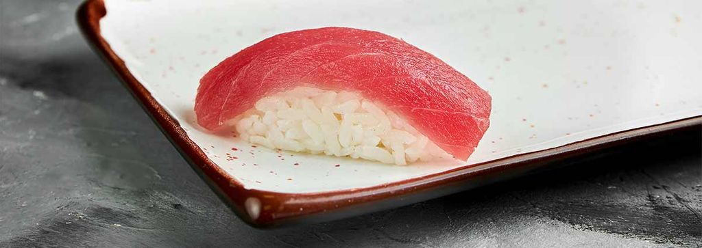 A piece of nigiri sushi with tuna on a white plate with a brown rim.