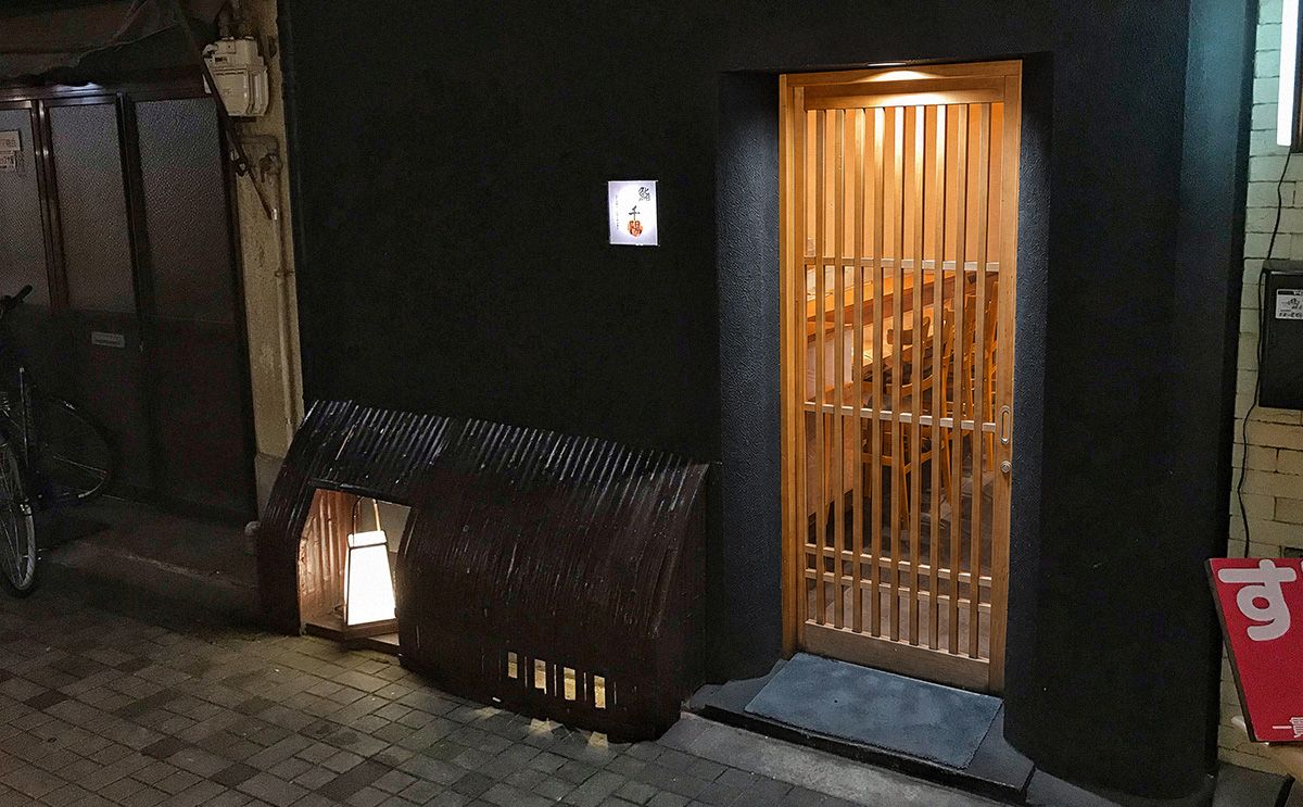 Außenansicht eines kleinen sushiya in Osaka bei Nacht, mit dunkler Fassade, Holzgittertür und warm beleuchtetem Eingang.