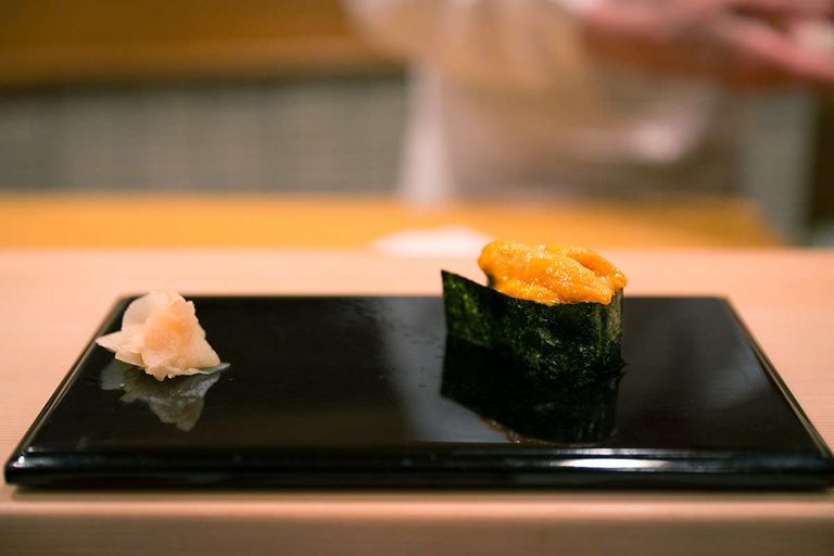 A piece of Uni Gunkan Sushi, consisting of sea urchin roe (uni) on sushi rice, wrapped in a strip of nori seaweed, served on a black plate with pickled ginger beside it.