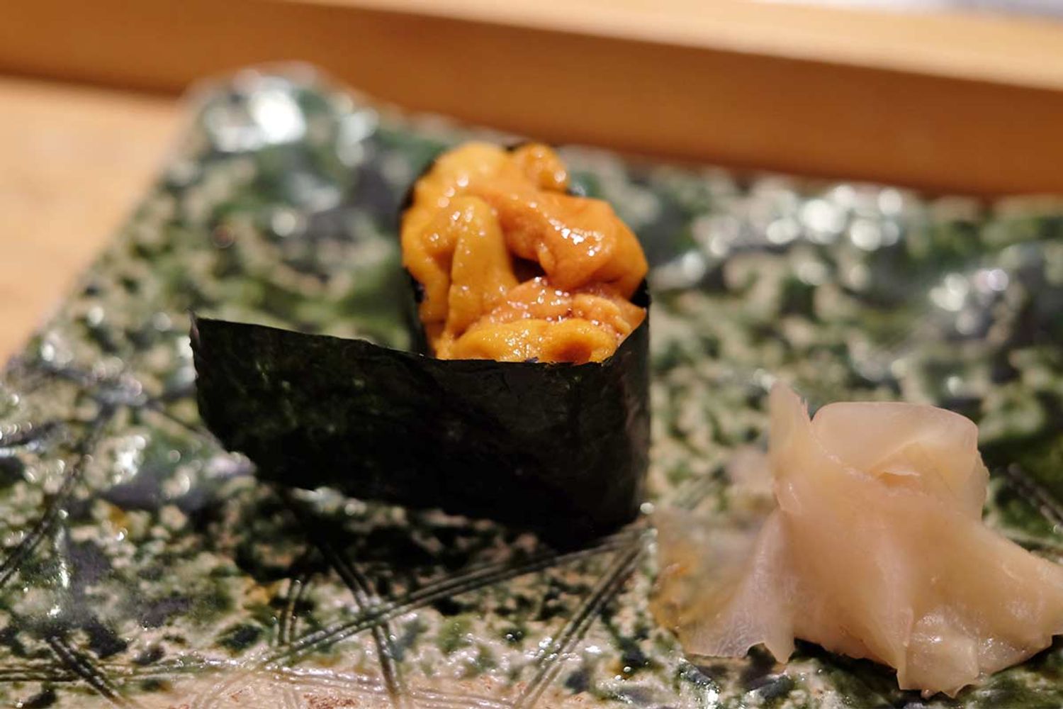 Ein Stück Uni Gunkan Sushi, bestehend aus Seeigel-Rogen (Uni) auf Sushi-Reis, umhüllt von einem Streifen Nori-Alge, serviert auf einem kunstvoll gestalteten Teller mit eingelegtem Ingwer daneben.
