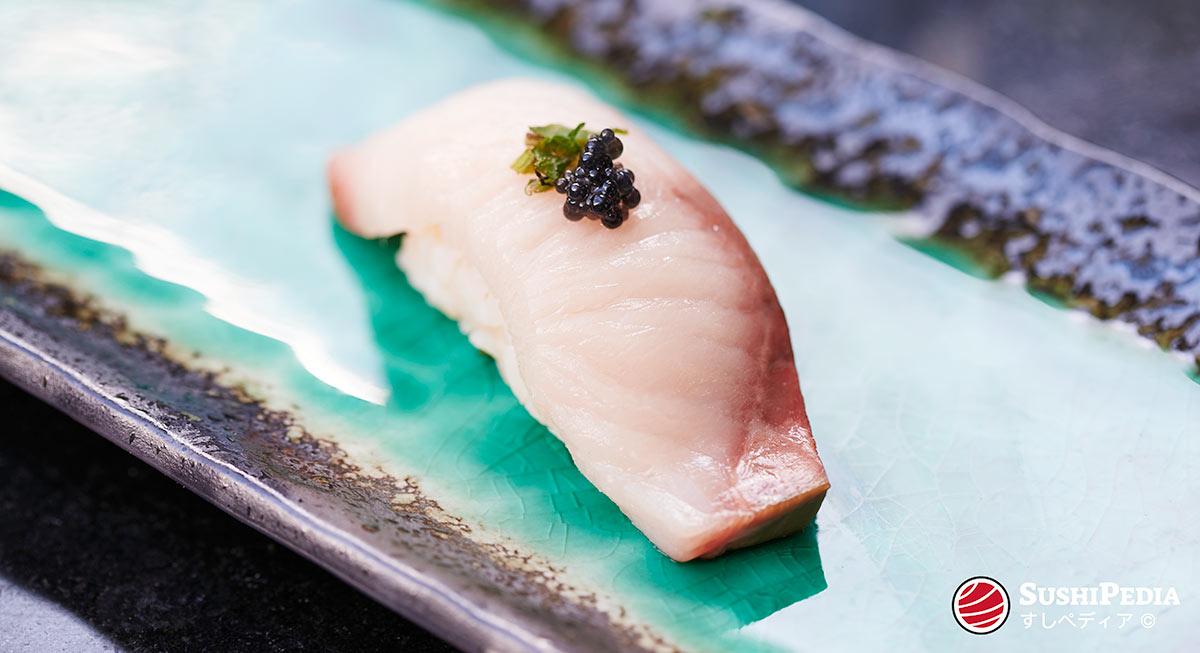Ein handgeformtes Sushi (jap. Nigiri) mit japanischen Amberjack bzw. Gelbschwanz (jap. Hamachi) liegt auf einem japanischen Keramikteller. Garniert ist es mit einem Topping aus mariniertem Shiso und Kaviarrogen.