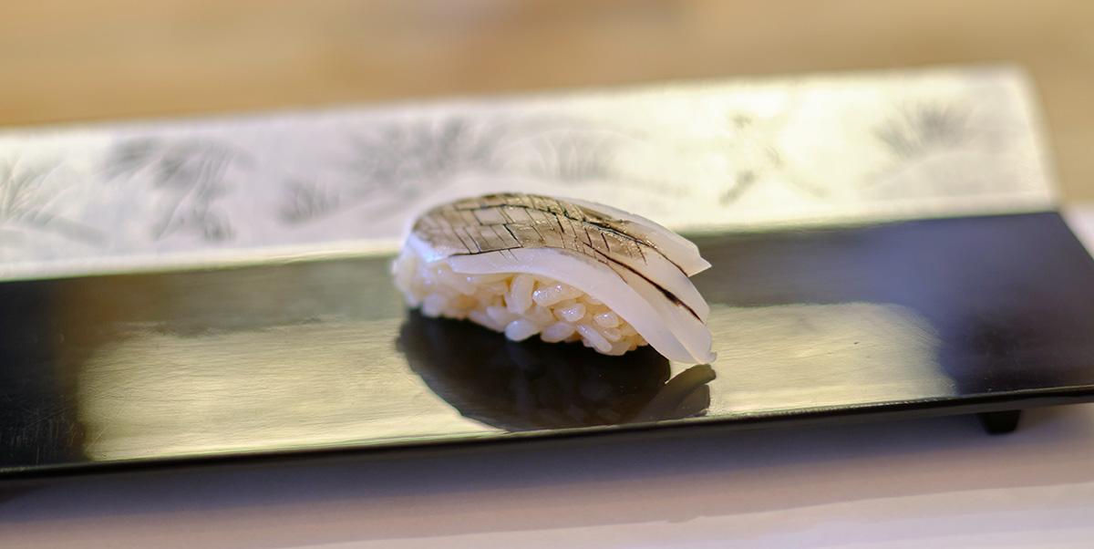 A piece of squid sushi nigiri (hand-shaped sushi) that has been coated with squid ink and is sitting on the counter of a sushi restaurant.